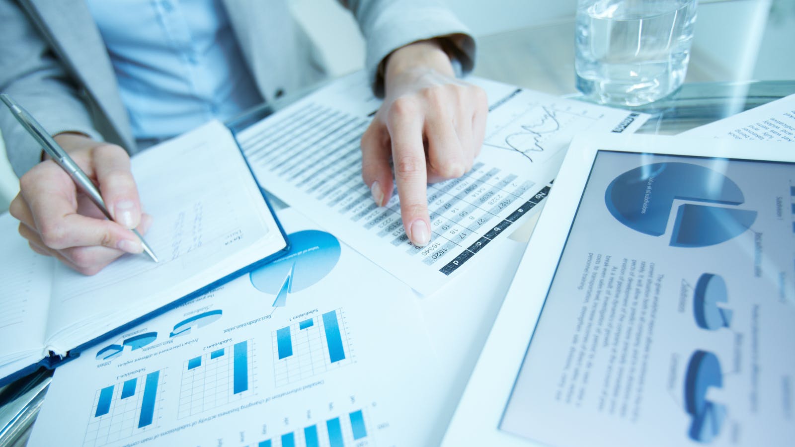 Businessman at desk examining graphic representations of data &mdash; tables and charts &mdash; on printed documents and a tablet.